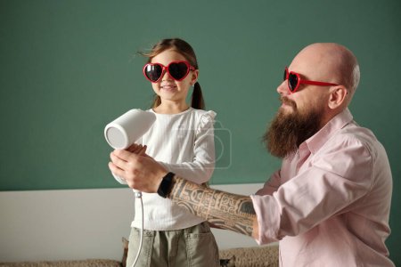 Photo for Cute happy girl standing in front of camera next to her bearded father in heartshaped sunglasses holding hairdryer by her face - Royalty Free Image