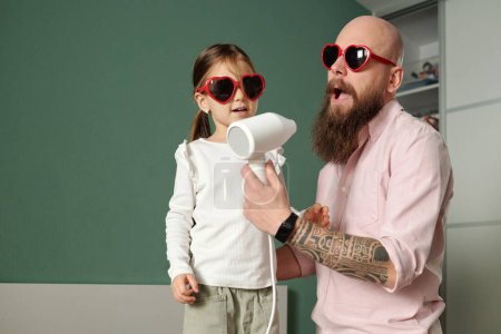 Photo for Adorable girl and her bald bearded young man in heartshaped sunglasses singing in hair dryer together while enjoying pastime - Royalty Free Image