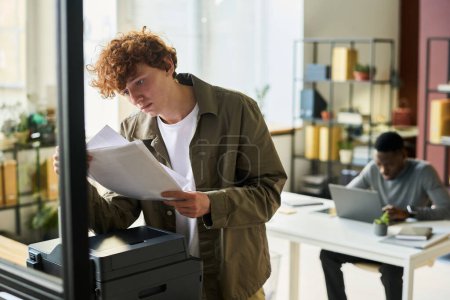 Foto de Joven trabajador de cuello blanco en ropa casual mirando los papeles en sus manos mientras está de pie frente a la máquina xerox en la oficina - Imagen libre de derechos