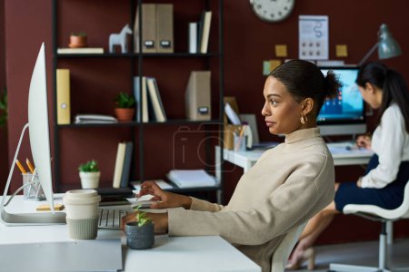 Foto de Vista lateral de la joven mujer de negocios afroamericana aburrida desplazándose a través de cosas en línea mientras mira la pantalla de la computadora - Imagen libre de derechos