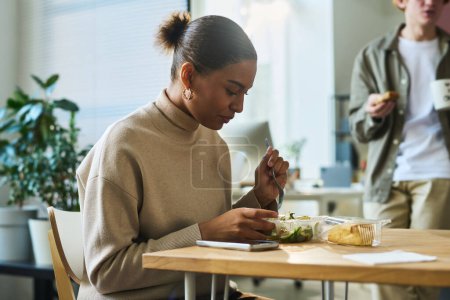Téléchargez les photos : Jeune ouvrière afro-américaine affamée assise à table à la pause déjeuner et ayant une salade végétarienne dans un récipient en plastique - en image libre de droit