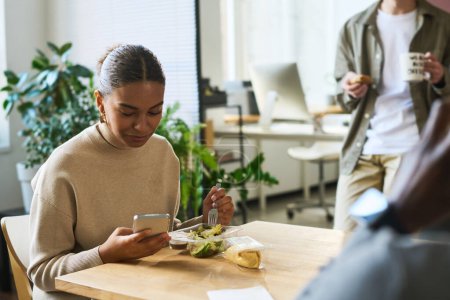 Photo for Young African American female office worker texting in smartphone or watching online video while having vegetarian salad by lunch - Royalty Free Image
