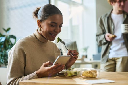 Téléchargez les photos : Heureuse jeune femme d'affaires afro-américaine regardant une vidéo sur smartphone et ayant une salade à la pause déjeuner au bureau - en image libre de droit