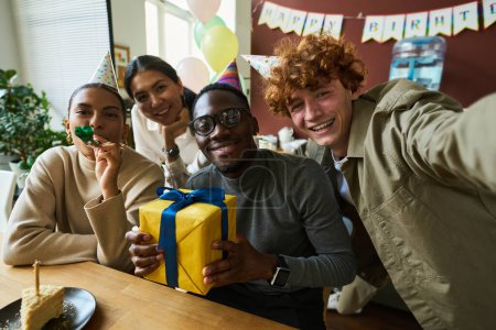 Foto de Jóvenes trabajadores de cuello blanco interculturales con ruidosos y regalo mirando a la cámara mientras alegre hombre de negocios tomando selfie cumpleaños - Imagen libre de derechos