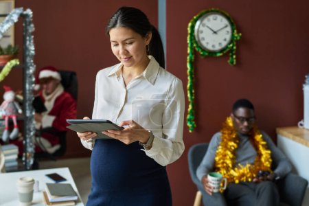 Téléchargez les photos : Jeune femme d'affaires enceinte asiatique regardant l'écran de la tablette tout en réseautant dans un bureau ouvert décoré pour Noël - en image libre de droit