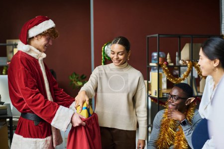Photo for Young smiling African American businesswoman taking giftbox out of red sack held by male colleague wearing Santa Claus costume - Royalty Free Image