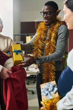 Foto de Feliz joven hombre de negocios negro con decoración de Navidad celebración envuelto paquete con regalo sobre saco rojo lleno de regalos para colegas - Imagen libre de derechos