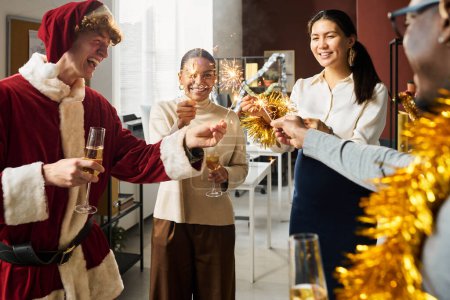Téléchargez les photos : Groupe de jeunes managers heureux avec des flûtes de champagne tenant des étincelles pendant la fête de Noël tout en profitant de la célébration - en image libre de droit