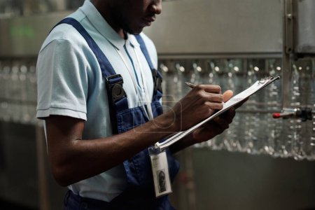 Téléchargez les photos : Jeune ingénieur afro-américain d'usine d'eau embouteillée remplissant sous forme de livraison tout en restant près de la chaîne de montage avec des bouteilles vides - en image libre de droit