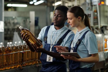 Téléchargez les photos : Jeunes ingénieurs interculturels du contrôle de la qualité regardant une bouteille en plastique contenant de la limonade carbonatée ou une autre boisson - en image libre de droit