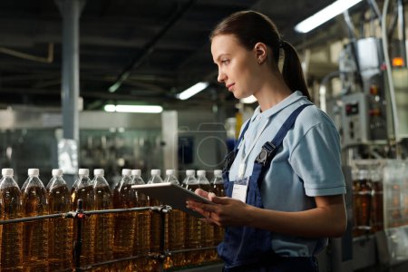Photo for Young quality inspector with tablet standing in front of assembly line with capped bottled drinks and examining plastic packaging - Royalty Free Image