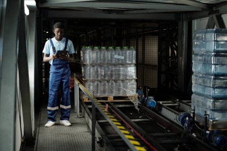 Téléchargez les photos : Jeune ouvrier afro-américain en salopette en utilisant une tablette tout en étant debout par une bande transporteuse avec des bouteilles en plastique avec des boissons - en image libre de droit