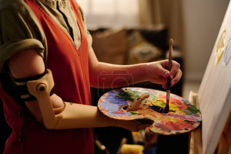 Téléchargez les photos : Main d'adolescente avec bras myoélectrique debout devant une œuvre d'art inachevée sur chevalet tout en créant une nouvelle peinture - en image libre de droit