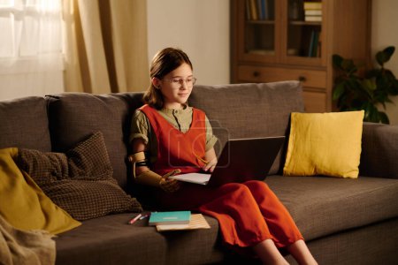 Photo for Clever schoolgirl with myoelectric arm sitting on sofa with cushions in living room and listening to teacher during online lesson - Royalty Free Image