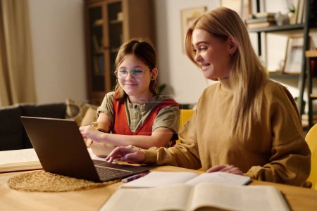 Photo for Smiling amputee girl looking at laptop screen while sitting by desk next to blond female tutor scrolling through points of lesson - Royalty Free Image