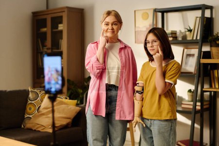 Téléchargez les photos : Jeune femme blonde en chemise rose et jeans bleus et sa jeune fille avec bras partiel gardant leurs index sous la zone des yeux - en image libre de droit
