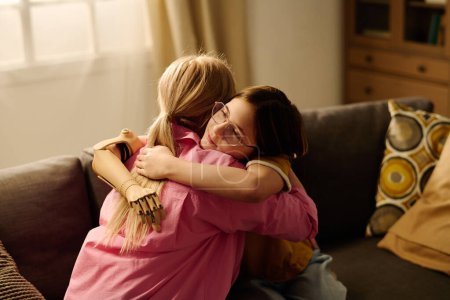 Téléchargez les photos : Jeune mère célibataire blonde et heureuse fille affectueuse avec la main myoélectrique assise dans l'étreinte sur le canapé dans le salon - en image libre de droit