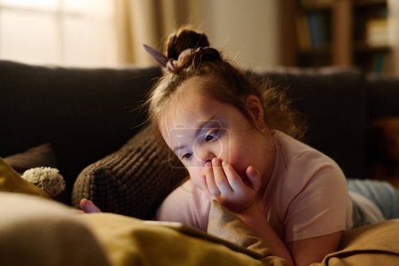 Foto de Adorable chica con síndrome de Down tumbado en el sofá y mantener la mano por la boca mientras ve dibujos animados en línea en la pantalla de la tableta - Imagen libre de derechos