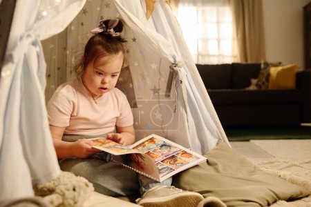 Téléchargez les photos : Douce fille avec le syndrome de Down assis sur le sol dans la tente et en regardant des photos dans le livre de bandes dessinées pour les enfants dans le salon spacieux - en image libre de droit