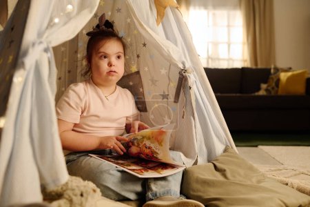 Photo for Cute child with Down syndrome sitting in tent in front of camera in living room with couch and holding book of comics for kids - Royalty Free Image