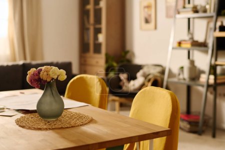 Photo for Part of wooden table with grey vase with bunch of flowers surrounded by two yellow chairs against interior of spacious living room - Royalty Free Image