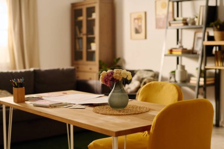 Téléchargez les photos : Table en bois avec papiers, crayons et vase gris avec des fleurs debout devant la caméra dans le centre du salon spacieux - en image libre de droit