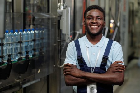 Téléchargez les photos : Jeune inspecteur afro-américain prospère en vêtements de travail regardant la caméra avec le sourire tout en étant debout par l'équipement industriel - en image libre de droit