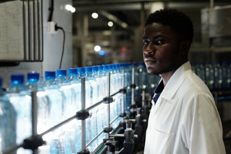 Téléchargez les photos : Jeune ingénieur afro-américain de contrôle de la qualité en blouse debout devant la chaîne de montage avec des boissons en bouteille bouchées - en image libre de droit