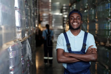Téléchargez les photos : Jeune ingénieur afro-américain réussi de l'entrepôt en vêtements de travail regardant la caméra contre d'énormes piles de paquets - en image libre de droit