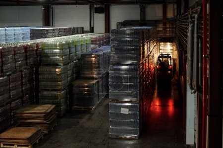 Photo for Forklift moving forward along aisle between huge stacks of packages with plastic bottles with mineral water or some other beverages - Royalty Free Image