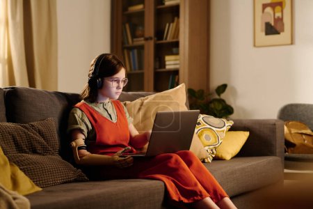 Téléchargez les photos : Adolescente handicapée assise sur un canapé dans le salon et regardant un écran d'ordinateur portable tout en regardant une vidéo ou un film en ligne - en image libre de droit