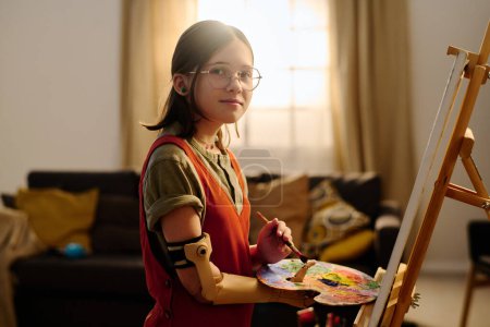 Téléchargez les photos : Pré-adolescente handicapée regardant la caméra tout en se tenant devant le chevalet dans le salon et en travaillant sur de nouvelles œuvres d'art créatives - en image libre de droit