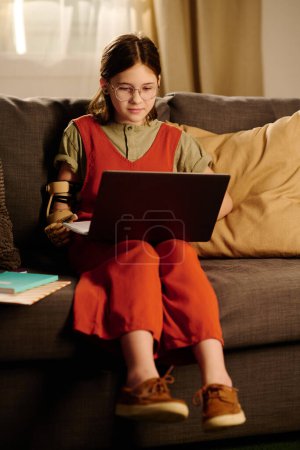 Téléchargez les photos : Écolière mignonne avec ordinateur portable sur ses genoux regardant l'écran tout en étant assis sur le canapé devant la caméra et ayant une leçon en ligne - en image libre de droit