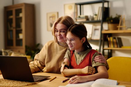 Téléchargez les photos : Écolière mignonne avec handicap et jeune femme regardant l'écran d'ordinateur portable tout en étant assis à côté du bureau l'un à côté de l'autre et en regardant la vidéo en ligne - en image libre de droit