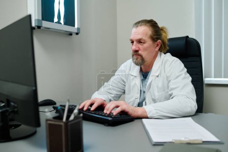 Foto de Médico serio con bata de laboratorio escribiendo en el teclado de la computadora de escritorio mientras consulta al paciente en línea por lugar de trabajo en el consultorio médico - Imagen libre de derechos