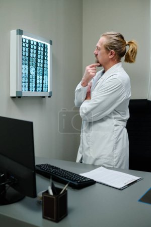Téléchargez les photos : Oncologue ou radiologue professionnel mature en blouse de laboratoire regardant ct scanner du cerveau du patient tout en analysant les résultats de l'examen - en image libre de droit