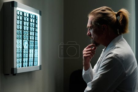 Téléchargez les photos : Radiologiste expérimenté coûteux avec la main par la barbe en regardant ct scanner du cerveau après examen du patient et en essayant de faire un diagnostic - en image libre de droit