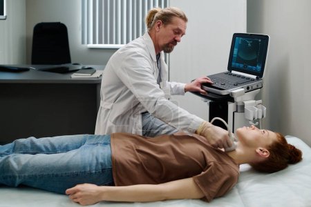 Téléchargez les photos : Endocrinologue mature en blouse mobile sonde ultrasonique sur le cou d'une jeune patiente assise près d'une machine à ultrasons - en image libre de droit
