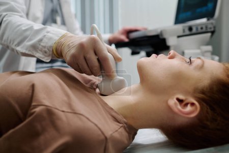 Téléchargez les photos : Jeune femme couchée au cabinet médical pendant la procédure d'échographie étant faite par endocrinologue dans des gants en caoutchouc avec sonde à ultrasons - en image libre de droit