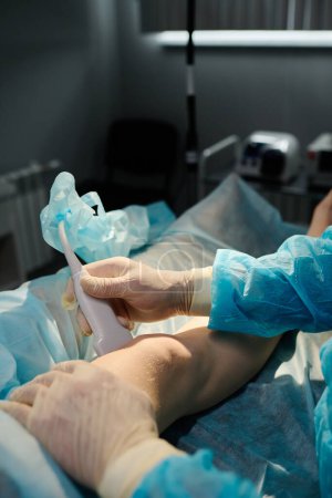 Téléchargez les photos : Mains du chirurgien en gants de caoutchouc protecteurs tenant la sonde près du genou de la jeune patiente allongée sur la table d'opération - en image libre de droit