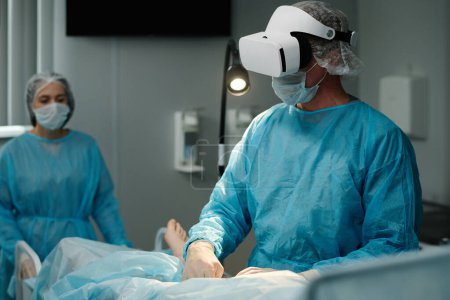 Photo for Mature surgeon in VR headset, scrubs, protective mask and gloves using medical equipment while performing surgical operation - Royalty Free Image