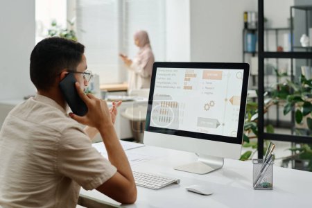 Téléchargez les photos : Jeune cols blancs occupés avec téléphone portable par oreille assis par lieu de travail et regardant l'écran d'ordinateur avec des données - en image libre de droit