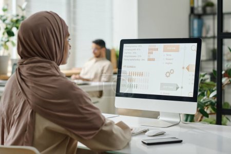 Photo for Young Muslim businesswoman in hijab sitting by desk in front of computer screen with statistic data and analyzing it against male coworker - Royalty Free Image