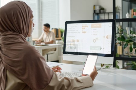 Photo for Young Muslim businesswoman in hijab holding smartphone with blank screen while sitting by workplace in front of desktop computer - Royalty Free Image