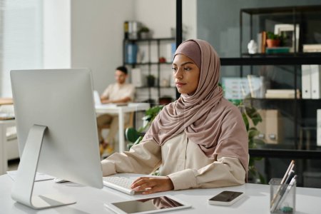 Téléchargez les photos : Jeune employée de bureau dans le hijab assise sur le lieu de travail devant un écran d'ordinateur et analysant les données dans un espace de coworking - en image libre de droit