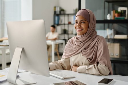 Téléchargez les photos : Joyeux jeune analyste ou courtier en hijab regardant l'écran d'ordinateur avec le sourire tout en étant assis par le lieu de travail et en analysant les données - en image libre de droit