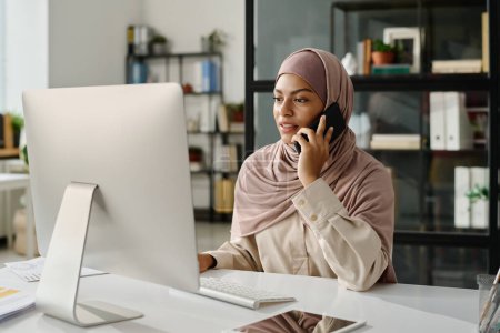 Téléchargez les photos : Jeune femme d'affaires musulmane confiante dans le hijab regardant l'écran d'ordinateur et parlant sur un téléphone portable dans un espace de coworking - en image libre de droit