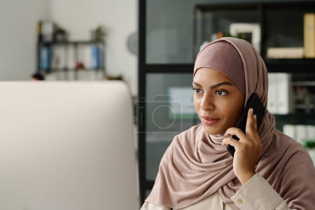 Téléchargez les photos : Ouvrier musulman confiant dans le hijab parler au demandeur sur un téléphone mobile et regarder l'écran d'ordinateur par lieu de travail - en image libre de droit