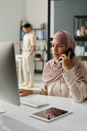 Photo for Young confident manager in hijab speaking on smartphone in front of desktop computer screen while sitting by workplace in office - Royalty Free Image
