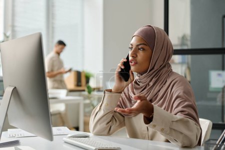 Téléchargez les photos : Jeune employée musulmane dans le hijab expliquant quelque chose au partenaire d'affaires au téléphone tout en étant assise sur le lieu de travail avec un ordinateur de bureau - en image libre de droit